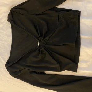 Svart croppad långärmad topp - Snygg svart croppad topp med lång ärm och ribbad struktur. Toppen har en cool v-ringning med snörning framtill och små pärlor som detalj i mitten. Perfekt för dig som gillar trendiga och unika plagg.