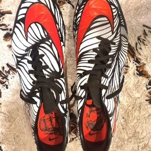 Nike Neymar jr fotbollsskor i svart, vitt och rött - Snygga Nike fotbollsskor i storlek 45 med ett unikt svartvitt grafiskt mönster och röda detaljer. Skorna har snörning, platt sula med dobbar och en stor röd Nike-swoosh på sidan. Perfekta för dig som vill sticka ut på planen.