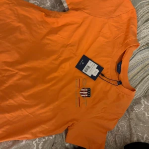 Orange t-shirt från GANT - Snygg orange t-shirt från GANT med broderad logga och emblem på bröstet. Klassisk rund hals och korta ärmar. Perfekt för dig som gillar sportig och clean stil. Materialet är mjuk bomull som känns skönt mot huden.( Priserna på alla varor kan diskuteras )