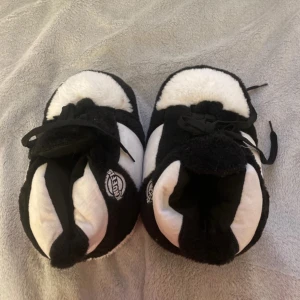 Svarta och vita fluffiga tofflor  KnuFFs - Supermysiga sneakers-inspirerade tofflor från KnuFFs i svart och vitt. De är gjorda i ett fluffigt syntetmaterial och har snörning framtill. Perfekta för att chilla hemma och hålla fötterna varma med en cool look.