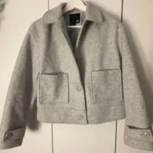 Ljusgrå ulljacka med stora fickor - Snygg ljusgrå jacka i ull med raka linjer och stora framfickor. Jackan har krage, knäppning framtill och coola detaljer vid ärmsluten. Perfekt för dig som gillar minimalistisk och clean stil.