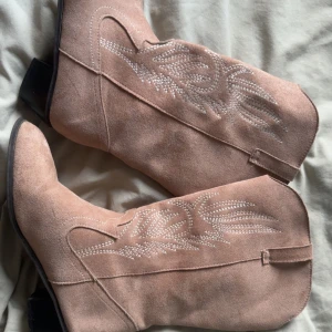 Beiga cowboy boots i mocka - Snygga beiga cowboy boots i mjuk mocka med klassiska broderade detaljer på skaftet. Boots med spetsig tå och låg klack, perfekta för att lyfta din outfit med western-vibe.