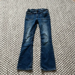 Blå bootcut jeans med fickdetaljer - Snygga blå jeans med bootcut passform och coola fickdetaljer både fram och bak. Jeansen har dubbla knappar i midjan och klassisk tvättad denimlook. Perfekta för dig som gillar en retrostil med lite utsvängda ben.