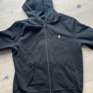 Svart hoodie från Polo Ralph Lauren - Svart hoodie från Polo Ralph Lauren i storlek L. Tröjan har huva med snörning, dragkedja framtill och två stora fickor. Klassisk vit broderad logga på bröstet. Perfekt för chill dagar och enkel att matcha med jeans eller joggers.