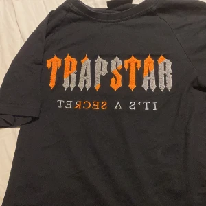 Svart Trapstar t-shirt med broderi - Svart t-shirt från Trapstar med fet broderad logga i orange och grått på bröstet. Texten 'IT'S A SECRET' är broderad under loggan. Klassisk rund hals och korta ärmar. Materialet är mjuk bomull, perfekt för en chill streetwear-look.