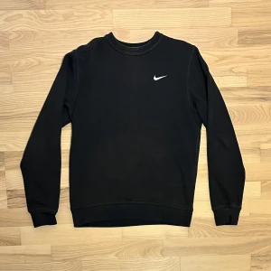 Nike sweatshirt svart - Svart crewneck sweatshirt från Nike med klassisk vit swoosh broderad på bröstet. Tröjan har rund hals, ribbade muddar och är tillverkad i mjuk bomullsmix. Perfekt för chill eller sportiga dagar, enkel och stilren design som funkar till allt.