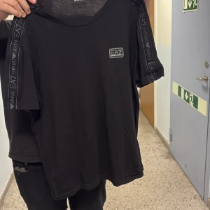 Svart EA7 Emporio Armani t-shirt - Svart t-shirt från EA7 Emporio Armani med korta ärmar och rund hals. Snygga logoband längs axlarna och ett stilrent EA7-märke på bröstet. Mjuk och skön bomullskvalitet som passar perfekt till jeans eller shorts. Enkel, clean och modern design. Helt oanvänd,vid frågor/bilder skriv privat 