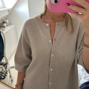 Beige kofta med knappar - Mysig beige kofta med rund hals och stora, ljusa knappar framtill. Tröjan har halvlånga ärmar och är stickad i ett mjukt material med ribbade detaljer vid hals, ärmslut och nederkant. 
