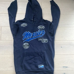 Mörkblå hoodie från Mercier - Snygg mörkblå hoodie från Mercier med stora broderade loggor och patchar framtill. Tröjan har huva med snörning och en klassisk känguruficka. Perfekt för dig som gillar streetwear och vill sticka ut med coola detaljer.