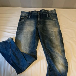 G-Star RAW blå jeans med slitningar - Säljer ett par blå jeans från G-Star RAW med snygga slitningar och tvättade detaljer. Jeansen har klassisk femficksdesign och tydliga logopatchar fram och bak. Perfekta för en avslappnad streetstil. 