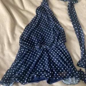 Marinblå prickig kortklänning  - Säljer en marinblå kortklänning från SHEIN med vita prickar och omlott-design. Klänningen har oneshoulder och knytband vid axeln för en snygg detalj. Materialet är mjuk polyester med lite stretch, perfekt för dig som gillar trendiga mönster och coola snitt.