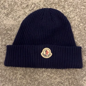 Mörkblå Moncler mössa i ull - Ribbstickad mörkblå mössa från Moncler med klassisk uppvikt kant och ikonisk logotyp framtill. Tillverkad i mjuk ull som håller dig varm och snygg. Perfekt accessoar för kyliga dagar och en stilren detalj till din outfit.Skicket är jätte bra om man vill ha mer bilder är det bara att skriva. Om du är intresserad bara skriv så löser vi