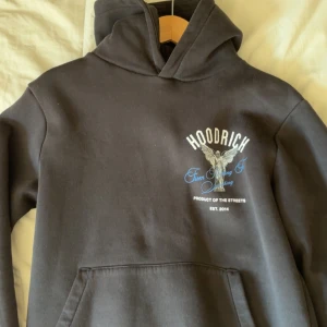 Svart Hoodie från Hoodrich med ängel - Svart hoodie från Hoodrich med stor ängelgrafik och texten 'From Nothing To Something' i blått på ryggen. Framsidan har Hoodrich-logga och mindre ängeltryck. Klassisk känguruficka och huva med broderad detalj. Mjuk bomullskvalitet, perfekt för streetwear.pris går att diskutera 