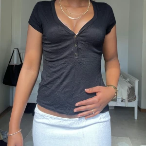 Svart v-ringad topp med knappar - Svart slim topp med korta ärmar och djup v-ringning framtill. Tre snygga knappar ger extra detalj och den sitter tajt mot kroppen. Perfekt att styla med smycken eller lager på lager. Mjuk bomullskänsla och enkel, clean look.