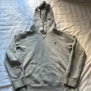 Grå hoodie från Polo Ralph Lauren - Säljer en grå hoodie från Polo Ralph Lauren med klassisk broderad logga på bröstet. Tröjan har huva, känguruficka och långa ärmar. Mjuk insida och stilren design som funkar till jeans eller joggers. Perfekt för chill dagar eller när du vill ha en enkel look.