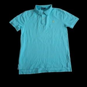 Turkos pikétröja från Polo Ralph Lauren - Snygg turkos pikétröja från Polo Ralph Lauren med klassisk krage och två knappar framtill. Tröjan har det ikoniska gula Polo-logot broderat på bröstet och är gjord i mjuk bomull. Perfekt för dig som gillar stilrena och sportiga plagg.