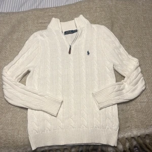 Vit stickad tröja Polo Ralph Lauren - Säljer en vit stickad tröja från Polo Ralph Lauren i storlek M. Tröjan har kabelstickat mönster, half zip och ribbade muddar. Klassisk broderad logga på bröstet. Perfekt för dig som gillar stilren och tidlös design.