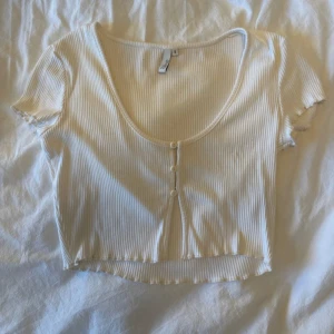 Vit ribbad croppad topp Gina Tricot - Superfin vit croppad topp från Gina Tricot med ribbad struktur och korta ärmar. Toppen har rundad urringning och knappar framtill. Perfekt till jeans eller kjol för en fräsch look.