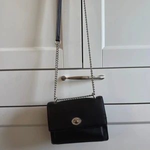Svart axelväska med kedja - Stilren svart axelväska i skinn med silverfärgad kedjerem och vridlås framtill. Väskan har en fyrkantig form och minimalistisk design som passar perfekt till din outfit. Enkel att bära crossbody eller över axeln.