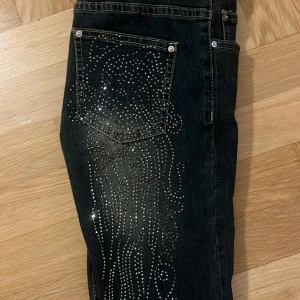 Low waist svarta  jeans med strass - Köpt här på Plick, säljer ett par svarta jeans med bootcut-modell och snygga strassdetaljer på bakfickorna och längs benen. Jeansen har en lätt tvättad look och glittrande mönster som ger extra edge. Perfekta för dig som vill sticka ut med din outfit. 