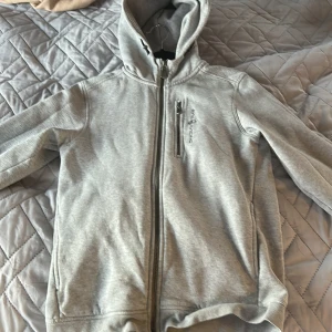 Grå hoodie från Sail Racing med dragkedja - Snygg grå hoodie från Sail Racing i storlek 170. Tröjan har hel dragkedja framtill, huva och en bröstficka med dragkedja. Perfekt för dig som gillar en clean och sportig stil. Mjuk insida och klassisk passform.