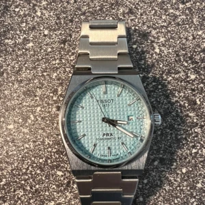 Tissot PRX Powermatic 80 blå klocka - Snygg Tissot PRX Powermatic 80 med ljusblå urtavla och stilrent mönster. Klockan har ett robust armband i silverfärgat stål och datumvisning vid klockan tre. Perfekt accessoar för dig som gillar klassisk design med modern touch.