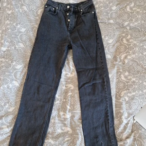 Mörkgrå jeans från A-DSGN, storlek S - Snygga mörkgrå jeans från A-DSGN i klassisk rak modell. Hög midja, fem fickor och knäppning med flera silverfärgade knappar framtill. Jeansen är tillverkade i kraftigt denimtyg och har en stilren look som passar till det mesta.