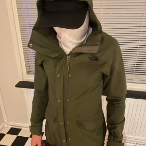 Grön vindjacka från The North Face - Snygg grön vindjacka från The North Face med huva och dragkedja framtill. Jackan har två fickor med lock och diskret logga på bröstet. Perfekt för blåsiga dagar och enkel att matcha med streetwear.