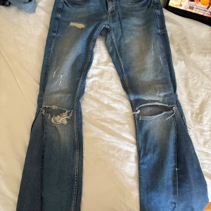 Skinny jeans omsydda till butcut från gina - Säljer ett par blå bootcut jeans från Gina. Jeansen har slitna detaljer och stora hål vid knäna för en edgy vibe. Klassisk femficksdesign och kontrastsömmar. Perfekt för dig som gillar en avslappnad och cool stil.