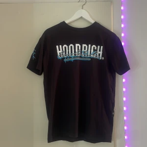 Svart HOODRICH t-shirt med tryck - Svart t-shirt från HOODRICH med stor vit och blå graffiti-inspirerad logga på bröstet och text på ryggen. Tryck även på ena ärmen. Rund hals och korta ärmar. Materialet känns som mjuk bomull, perfekt för streetwear-stil.