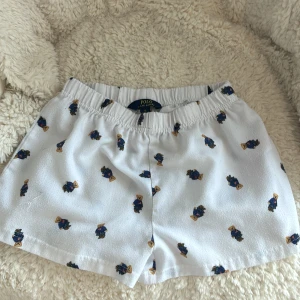 Vita pyjamas shorts med björnmönster Polo Ralph Lauren - Säljer ett par vita shorts från Polo Ralph Lauren med gulligt björnmönster över hela tyget. De har elastisk midja och är gjorda i mjuk bomull, perfekta för chill dagar. Passar dig som gillar klassisk stil med en twist.