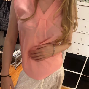 Ljusrosa ärmlös blus med v-ringning - Supersöt ljusrosa blus med v-ringad hals och snygg vikt detalj framtill. Modellen är ärmlös och toppen har en mjuk, flowy känsla som passar perfekt till sommaren. Enkel att matcha med både jeans och kjol för en fin look. Sparsam använd, o storlek M 