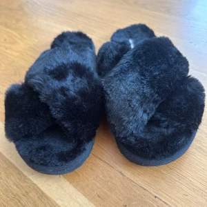 Svarta fluffiga sandaler från Even&Odd - Mysiga svarta sandaler från Even&Odd med korslagda band i mjuk fuskpäls. Sulan är platt och har en räfflad undersida för extra grepp. Perfekta för att chilla hemma eller när du vill ha något extra cozy på fötterna.