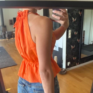 Orange ärmlös linnetopp med knyt - Säljer en superfin orange linnetopp med halterneck och knyt i nacken. Toppen har en luftig, volangig nederdel och smockad midja som ger en snygg siluett. Perfekt för varma dagar och enkel att styla med jeans eller kjol.