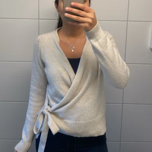 Beige omlottkofta med knyt - Mysig beige omlottkofta med knyt i sidan och långa ärmar. Koftan är stickad och har en mjuk känsla, perfekt att slänga på över en topp. Passar dig som gillar enkel och clean stil med lite extra detalj vid midjan.