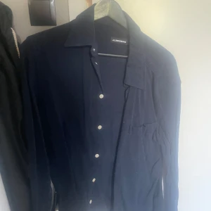  J.Lindeberg Mörkblå skjorta  - Nyrpis 1400kr. Snygg mörkblå skjorta från J.Lindeberg i mjuk subtil manchester. Klassisk krage, snygga vita knappar och en bröstficka. Långärmad och med normal passform, perfekt att bära öppen över t-shirt. Stilren och enkel att matcha med jeans eller chinos. Passar medium och smal. 