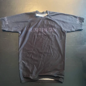 Svart Askari rashguard/compresionshirt  - En Svart OG rashguard/compresion shirt från Askari med texten 'WE OWN THE CAGE' tryckt på bröstet. Korta ärmar och en rund hals. Man får +10% guilliotin 