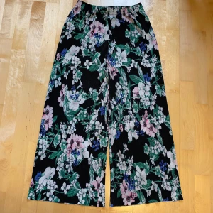 Blommiga vida byxor Gina Tricot XS - Säljer ett par svarta vida byxor från Gina Tricot i storlek XS med färgglatt blommönster i grönt, blått, rosa och vitt. Byxorna har resår i midjan och är tillverkade i mjuk polyester. Perfekta för dig som gillar statement-plagg och bekväm passform.