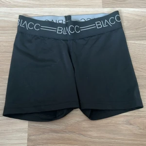 Svarta träningsshorts från BLACC - Svarta träningsshorts från BLACC med bred elastisk midja och logga i vitt. Sitter tight och är gjorda i mjukt, stretchigt material med 85% polyester och 15% elastan. Perfekta för gymmet eller sport, riktigt sköna och flexibla.