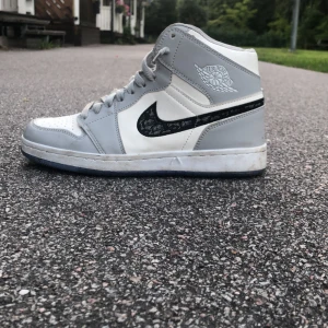 Nike Air Jordan 1 x Dior sneakers - Exklusiva Nike Air Jordan 1 x Dior sneakers i grått och vitt med svart Dior-mönstrad swoosh. Hög modell med snörning och rund tå. Sulan har genomskinliga blå detaljer med Dior-logga och Air Jordan-vinge. Skorna är tillverkade i skinn och har en riktigt clean och lyxig vibe.