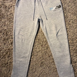 Grå mjukisbyxor från Trapstar - Snygga ljusgrå mjukisbyxor från Trapstar med broderad logga i blått, vitt och svart på ena benet. Byxorna har dragsko i midjan, fickor på sidorna och dragkedja vid benslut. Perfekta för chill eller streetwear.