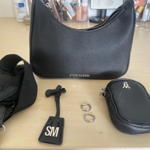 Steve Madden axelväska  - Snygg svart axelväska från Steve Madden i syntetmaterial med silvriga metalldetaljer och dragkedja. Ringarna som höll fast kedjan till väskan har gått av men det går att fixa (därav priset) Utöver det är väskan som ny! Inga fläck eller slitage. Väskan har även avtagbar rem, extra liten pouch med logga och SM-tag.