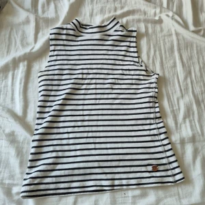 Randig ärmlös topp från Smiley - Vit och svart ribbad ärmlös topp med smala horisontella ränder och liten mockadetalj med smiley-logga nertill. Högre halsringning och mjukt, stretchigt material som sitter snyggt. Perfekt för dig som gillar enkel men cool stil.