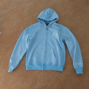 Ljusblå hoodiejacka  - Ljusblå hoodiejacka med broderade drakar på huvan och vita tryckdetaljer på bröst, rygg och ärm. Jackan har dragkedja framtill, känguruficka och ribbade muddar. Perfekt för dig som gillar streetwear och snygga detaljer. Priset är inte hugget I sten 🪨