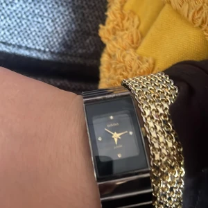 Rado Jubile herrklocka i silver - Stilren Rado Jubile herrklocka med rektangulär svart urtavla, guldfärgade visare och markeringar. Armband i blankt silverfärgat rostfritt stål. Klassisk och elegant design som sticker ut med sin unika form och lyxiga detaljer.