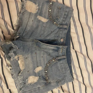 Blå jeansshorts med pärlor och nitar - Snygga blå jeansshorts med slitna detaljer, fransig kant och dekorerade med pärlor och nitar vid fickorna. Shortsen har klassisk femficksdesign och dragkedja framtill. Perfekt för dig som gillar en edgy och trendig stil. Passar xs-s