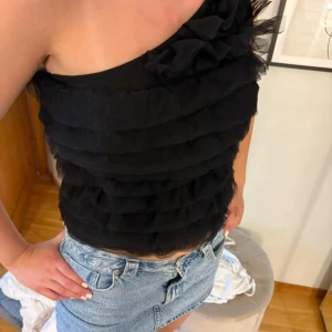 Svart oneshoulder topp från Stockh LM - Supersnygg svart oneshoulder topp från Stockh LM Studio med volangiga lager i tunn mesh. Toppen har en axel och cool ruffle-detalj upptill som ger extra edge. Perfekt att styla med jeans eller kjol för en trendig look.