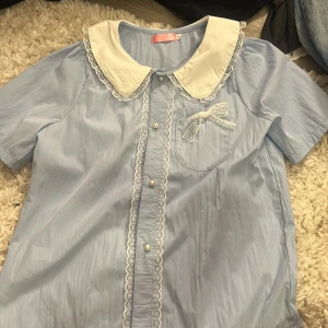 Ljusblå söt skjorta med spetsdetaljer - Supersöt ljusblå skjorta från Fei Xiang med vit krage och spetskant. Knappar framtill med pärlimitation och en liten spetsrosett på bröstfickan. Kortärmad och med mjukt, tunt material som känns skönt mot huden. Perfekt för dig som gillar gulliga detaljer.