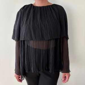 Svart plisserad blus med transparenta ärmar - Säljer en svart plisserad blus med transparenta, långärmade ärmar och lös passform. Blusen har ett lager över axlarna som ger en cool cape-effekt och är tillverkad i ett lätt, flowy material. Perfekt för dig som gillar unika detaljer och stilren look.