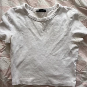 Vit croppad t-shirt från Zara - En enkel och fräsch vit t-shirt från Zara i croppad modell. T-shirten har rund hals och korta ärmar, perfekt för en clean och chill stil. Materialet är mjuk bomull som känns skönt mot huden. Passar dig som gillar basic och stilrent.